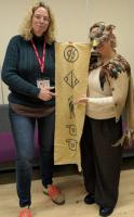 Annick and Ursula, dressed in a hawk costume, both smiling at the camera holding a fabric scroll with five Luwian hieroglyphs.
