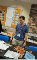 Gustavo standing behind a table with balls of clay, wooden styli, and a map of ancient Mesopotamia. The sign says 'Write Like Babylon'
