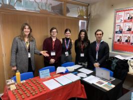 Five people behind a table with a sign saying 'Write Like A Mycenian Scribe', with clay balls, wooden styli and A4 information sheets.